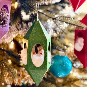 May include: A green paper Christmas ornament shaped like a house with a white snowy scene inside. The ornament has a glitter finish and a small deer and a tree inside.