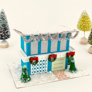 Puede incluir: Una casa de papel azul con un techo blanco y nieve en la parte superior. La casa tiene una valla blanca, una puerta verde y una corona en la puerta. Hay tres árboles pequeños frente a la casa y un camino que lleva a la puerta.