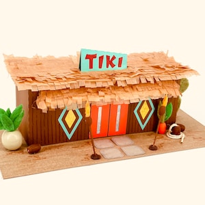 Op de afbeelding: Een bruine kartonnen tiki-hut met een rieten dak, oranje deuren en een bord met de tekst "TIKI". De hut heeft een blauw en geel ruitpatroon op de muren en is versierd met palmbomen en fakkels.