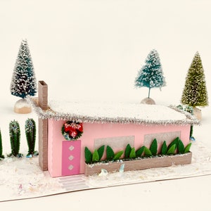 Puede incluir: Una casa de papel rosa con un techo blanco y una corona roja y verde en la puerta. La casa está rodeada de pequeños árboles verdes y una base blanca nevada.
