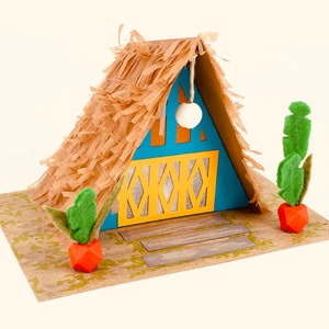 Puede incluir: Una casa en miniatura de papel con techo de paja, paredes azules y amarillas y una pequeña luz blanca colgando del techo. La casa está rodeada de plantas verdes y se asienta sobre una base de papel marrón.