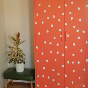 May include: A two-door metal locker with a coral orange finish and white polka dots. The locker has two doors with a handle on each door. The locker is standing on a wooden floor.