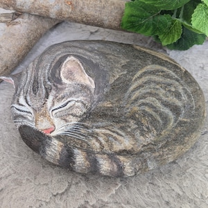 Puede incluir: Una piedra de río lisa y ovalada pintada a mano con la imagen detallada de un gato atigrado durmiendo. El gato presenta un pelaje a rayas grises y marrones, ojos cerrados y una cola enroscada, capturando al felino en posición de descanso.