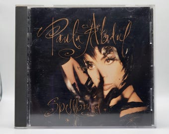 May include: A Paula Abdul Spellbound music CD in a clear plastic jewel case. The cover features a dark, moody portrait of the artist with black feathers, accented by copper-colored cursive text that reads Paula Abdul and Spellbound.