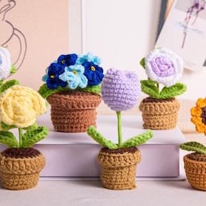 May include: A collection of handmade crochet flowers in small brown pots. The flowers include a blue and white rose, a yellow rose, blue flowers, a purple tulip, a white and purple rose, and a sunflower. The flowers have green leaves.