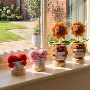 May include: Two crocheted heart figures in red and pink, and two crocheted sunflower figures in brown pots, all mounted on small wooden bases. Each figure holds a small white card with printed text. The items are arranged on a window sill.