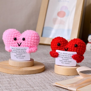 May include: Two crocheted heart figures, one pink and one red, each with black bead eyes and a stitched mouth. They are mounted on round wooden bases and hold a white card that reads: Mom, thank you for raising me to be a strong, independent woman.