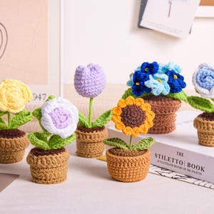 Puede incluir: Una colección de flores de ganchillo hechas a mano en pequeñas macetas marrones. Las flores incluyen una rosa amarilla, una flor blanca y morada, un tulipán morado, un girasol, flores azules y una rosa blanca y azul. El libro "The Stiletto Book" es visible.