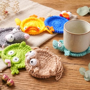 May include: A collection of six round, crocheted animal coasters in various colors including yellow, blue, gray, green, and tan. Each coaster features stitched facial details like eyes and limbs. A beige ceramic mug sits on the teal coaster.