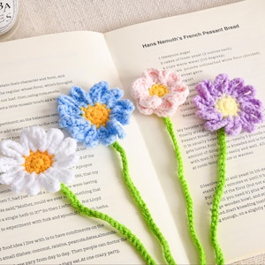 May include: Four handmade crochet flower bookmarks in white, blue, pink, and purple, each with a yellow centre and green stem. The bookmarks are placed on an open book with text.