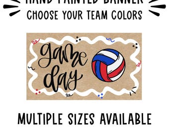 May include: A rectangular hand-painted banner on brown paper featuring the black text "game day" in a cursive script, a red, white, and blue volleyball graphic, and a white wavy border with small colorful confetti accents and text above and below.