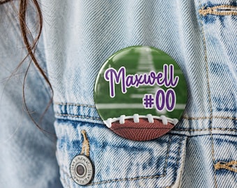 May include: A circular pinback button featuring a green football field background with white yard lines. The text Maxwell #00 is printed in purple with a white outline. A brown football with white laces is visible at the bottom of the button.