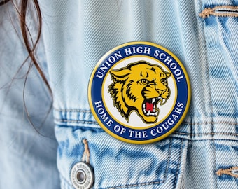 May include: A circular pinback button featuring a yellow cougar head graphic on a white background, surrounded by a blue border with the white text Union High School Home of the Cougars, attached to a light blue denim fabric garment.