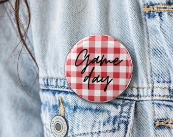 May include: A circular pinback button featuring a red and white checkered gingham pattern. The text "Game day" is printed across the center in a black cursive script font. The button is attached to a blue denim fabric material.