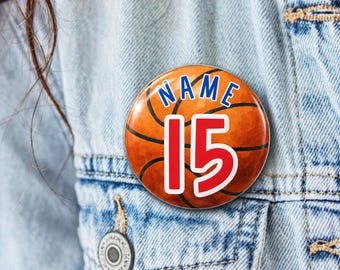 May include: A circular pinback button featuring a basketball graphic with black lines. The word NAME is printed in blue at the top, and the number 15 is displayed in large red digits with a white outline in the center of the basketball.