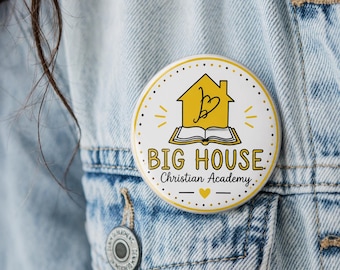 May include: A circular pinback button featuring a white background with a yellow house icon above an open book. The text reads BIG HOUSE Christian Academy in black and yellow lettering. A small yellow heart is centered below the text.
