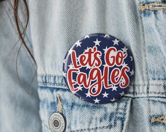 May include: A circular pinback button featuring a dark blue background with a repeating pattern of white stars. The text Let's Go Eagles is printed in a bold, red, cursive-style font across the center of the button, pinned to a denim jacket.