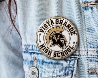 May include: A circular pinback button featuring a black and gold Spartan helmet logo in the center. The text VISTA GRANDE HIGH SCHOOL is printed in black capital letters along the white border, framed by a gold circular outline on a denim fabric.