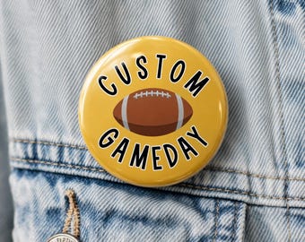 May include: A circular pinback button with a mustard yellow background featuring a brown American football graphic in the center. The text "CUSTOM GAMEDAY" is printed in bold black capital letters, arching above and below the football graphic.