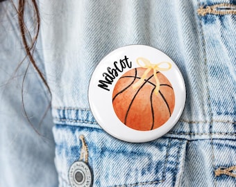 May include: A round white pinback button featuring a watercolor-style orange basketball with black lines and a small yellow ribbon bow on top. The word Mascot is printed in black cursive script to the left of the basketball graphic.
