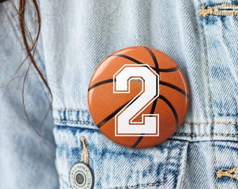 May include: A round pinback button featuring a textured orange basketball design with black lines and a large white number 2 printed in the center. The button is attached to a light blue denim fabric material with visible stitching and a metal button.