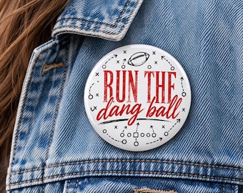 May include: A circular pinback button featuring a white background with black football play diagrams and a small football icon. The text "RUN THE dang ball" is printed in a distressed red font across the center of the button design.
