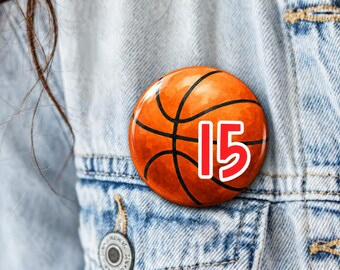 May include: A circular pinback button featuring a basketball graphic with black lines and the number 15 printed in bold red with a white outline. The button is attached to a light blue denim fabric surface with visible stitching.