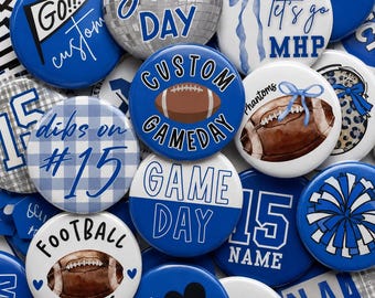 May include: A collection of round pin-back buttons featuring blue and white sports-themed designs. Graphics include footballs, pom-poms, a disco ball, and text such as Game Day, Dibs on #15, Football Mom #15, and Let's go MHP. The buttons vary in color.