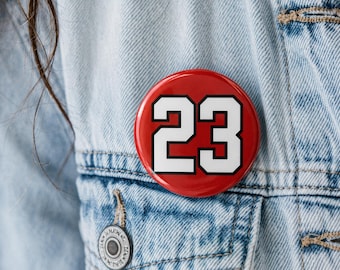 May include: A circular pinback button featuring a vibrant red background with the number 23 printed in a bold white font with a thick black outline. The button is attached to a light blue denim jacket with visible stitching and a metal button detail.
