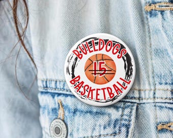 May include: A round pinback button featuring a central orange basketball graphic with the number 15 in white. The text BULLDOGS BASKETBALL is printed in red around the basketball, framed by a black circular brushstroke design.