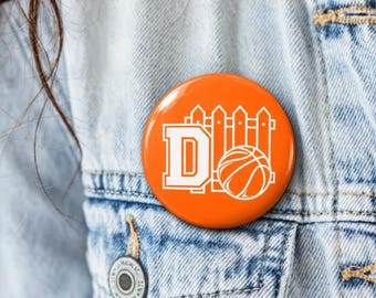 May include: A round orange pin-back button featuring a white graphic design of a picket fence, the letter D in a block font, and a basketball. The button is attached to a light blue denim jacket with visible stitching and a metal button.