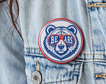 May include: A circular pinback button featuring a blue and white illustration of a bear wearing red-rimmed sunglasses. The text "BE ARS" is printed in white across the lenses of the sunglasses. The button has a red border and a white background.