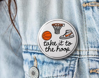 May include: A circular white pinback button featuring a black line drawing of a basketball hoop, a basketball, and a high-top sneaker with orange accents. The text "take it to the hoop" is printed in black cursive script below the illustrations.