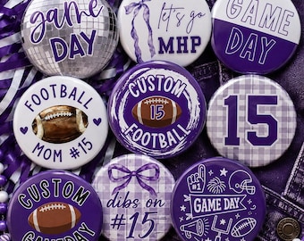 May include: A collection of nine round pinback buttons featuring purple and white designs. Graphics include a disco ball with text game DAY, a bow with let's go MHP, a football with FOOTBALL MOM #15, and various game day themed illustrations.