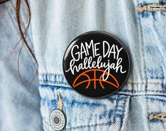 May include: A circular pinback button with a black background featuring the white text "GAME DAY" above the white text "hallelujah" and an orange graphic of a basketball. The button is attached to a light blue denim fabric.