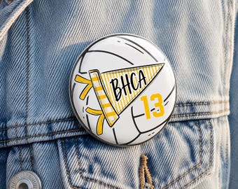 May include: A round pinback button featuring a white volleyball graphic with black lines. Centered on the ball is a yellow and white striped pennant with the black text BHCA and the yellow number 13 positioned to the right of the pennant.