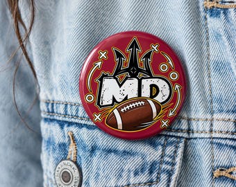 May include: A circular pinback button featuring a maroon background with a black trident icon above the white text MD. A brown American football with white laces is positioned below the letters, surrounded by white graphic arrows and cross symbols.