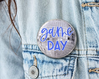 May include: A circular pinback button featuring a silver disco ball pattern with the text "game DAY" written in a bold, blue, sans-serif font with a white outline, centered on the reflective surface of the round accessory.