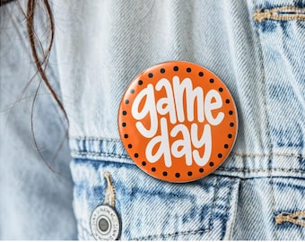 May include: A circular pinback button featuring a vibrant orange background with a black polka dot border. The words "game day" are written in a bold, white, rounded script font across the center of the button, creating a clear and legible design.