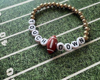 May include: A beaded bracelet featuring metallic gold-toned spherical beads and white circular beads with black lettering that spells out TOUCH DOWN. A small, brown, oval-shaped football-themed bead is positioned between the two words.
