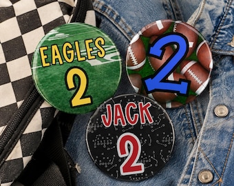 May include: Three circular pinback buttons featuring sports themes. One is green with the text EAGLES and the number 2. Another shows brown footballs on green grass with a blue number 2. The third is black with white play diagrams, the name JACK, and a red number 2.