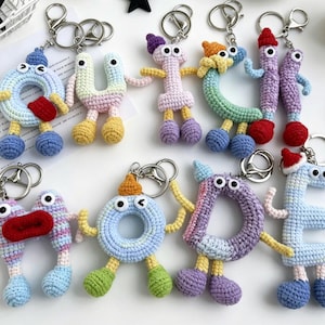 May include: A collection of colorful, handmade crochet keychains shaped like letters and numbers. Each keychain features a unique design with googly eyes, arms, and legs. The keychains are attached to silver metal rings.