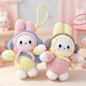 Peut inclure: Deux porte-clés en peluche en forme de lapin aux couleurs pastel. L'un a des oreilles roses et une tenue bleue et jaune, l'autre a des oreilles jaunes et une tenue rose et grise. Les deux ont des yeux noirs et des joues roses.