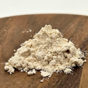 以下が含まれることがあります： 木製の表面に置かれた、食品成分またはサプリメントと思われる、薄いベージュ色の粉末の山をクローズアップで撮影。粉末はきめ細かい粒状の質感で、形はわずかに不均一です。背景はぼやけた木製の表面。