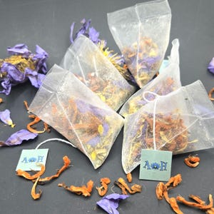 Puede incluir: Primer plano de varias bolsitas de té en forma de pirámide llenas de hierbas y flores secas. Las bolsas transparentes revelan una mezcla de plantas naranjas y moradas. Cada bolsa tiene una pequeña etiqueta con las letras "A H".