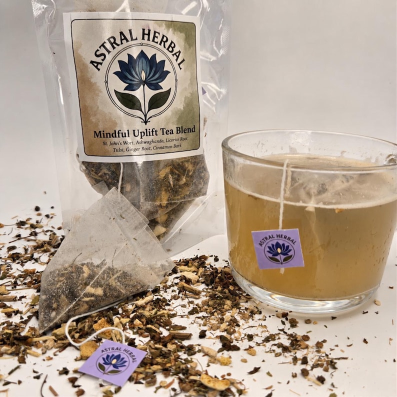 May include: A clear glass of tea with a tea bag and a bag of tea blend. The tea bag has a purple label with a blue lotus flower and the words "Astral Herbal". The bag of tea blend has a label that reads "Mindful Uplift Tea Blend".