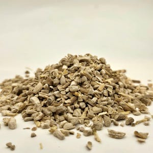 May include: A pile of chopped, light brown, dried plant material. The pieces vary in size and shape, with a mix of textures. The background is a plain, off-white color, providing a neutral backdrop for the product.