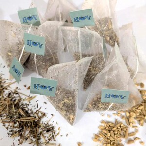 May include: A collection of pyramid-shaped tea bags filled with loose tea leaves. Each bag has a small, light blue tag with the letters "A H" and a floral design. Dried herbs and spices are scattered around the tea bags, suggesting a natural tea blend.