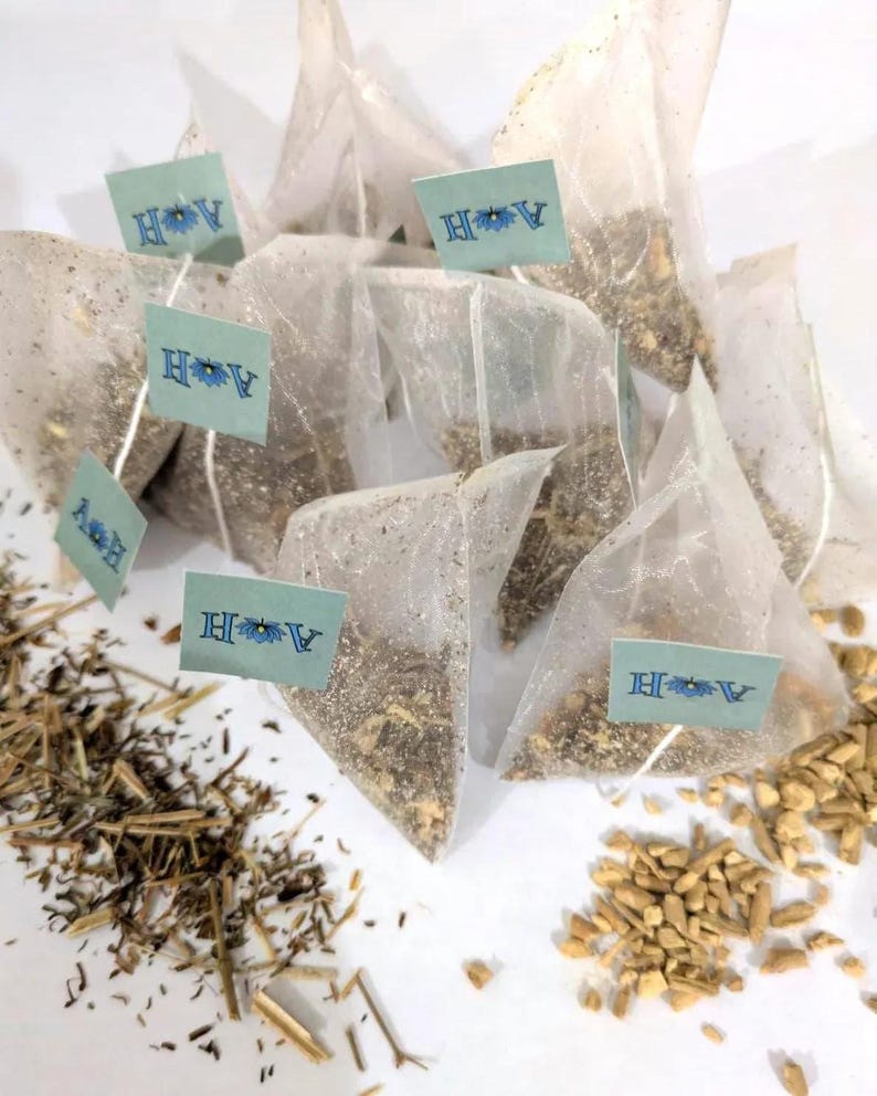 May include: A collection of pyramid-shaped tea bags filled with loose tea leaves. Each bag has a small, light blue tag with the letters "A H" and a floral design. Dried herbs and spices are scattered around the tea bags, suggesting a natural tea blend.