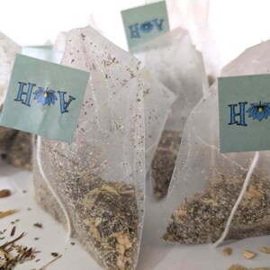 May include: Several tea bags filled with loose leaf tea are arranged on a white surface. Each tea bag is made of a sheer, white material and has a small, light blue tag attached. The tags display the letters 'A' and 'H' for Astral Herbal with a blue lotus flower in between. The tea bags are surrounded by loose tea leaves and small pieces of ginger.